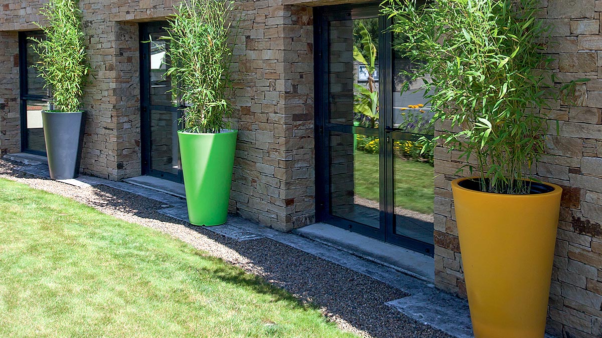 Trois demi-pots de fleurs plastique vert et orange collés contre une façade de maison de part et d'autres des fenêtres, pelouse et soleil