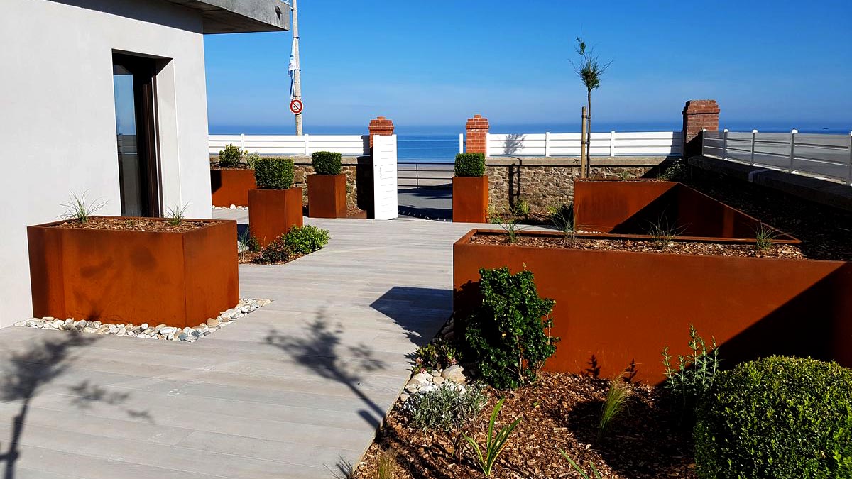 photo montrant un jardin sur fond de ciel bleu, une maison à gauche avec des bacs de jardin en corten à droite et gauche de l'image