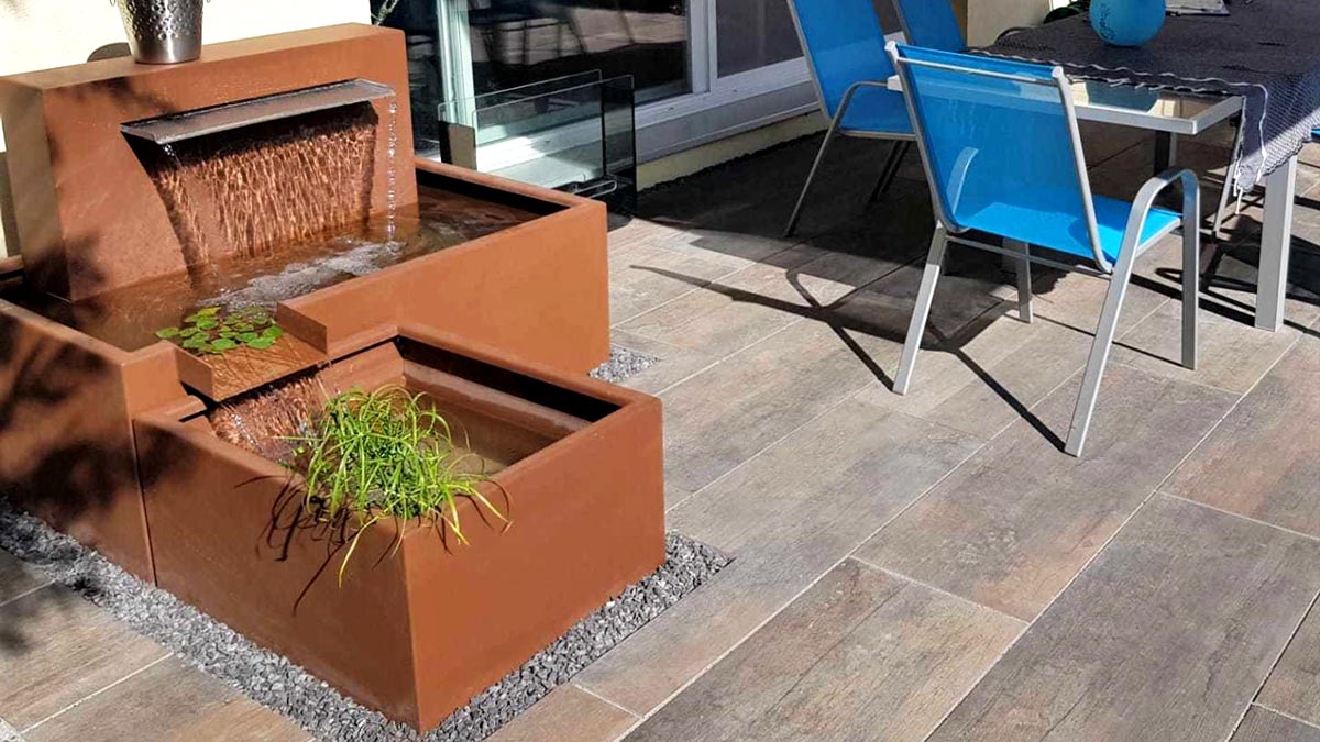 Fontaine de jardin en acier corten à gauche de l'image avec de l'eau qui coule, terrasse minérale, chaise bleue à droite