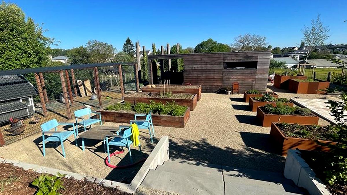 Jardin avec une allée centrale en graviers, des carrés potagers de part et d'autre, des chaises bleues à gauche, du soleil